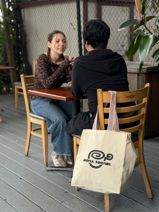 Two people sitting at a table outdoors with a tote bag featuring the A'Roma Roasters logo.